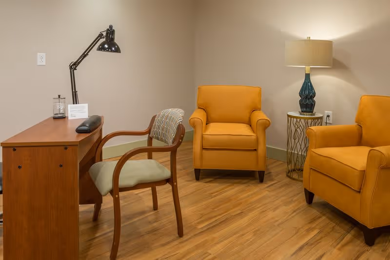 A cozy room with two mustard yellow armchairs, a small round side table with a blue lamp, and a wooden desk with a black adjustable desk lamp and a wooden chair with a patterned cushion. The floor is wooden and the walls are painted beige.