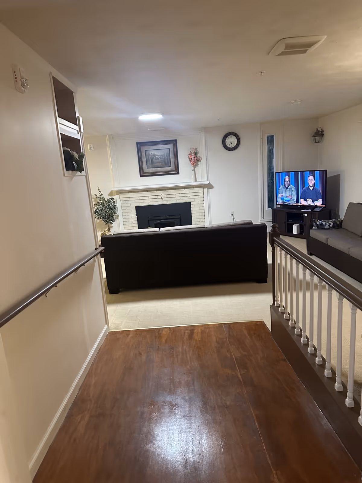 Interior view of a living room area in a senior living facility. The room features a dark sofa facing a white brick fireplace with a framed picture and a vase with flowers on the mantel. To the right, there is a TV on a stand showing two men, and another gray sofa with a decorative pillow. The floor transitions from wooden flooring in the foreground to carpet in the living area. A clock is mounted on the wall above the fireplace.