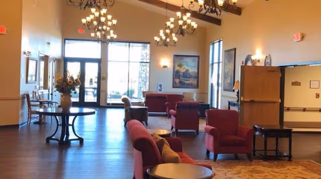 A spacious living room area with wooden flooring, multiple red upholstered armchairs and sofas, round tables, chandeliers hanging from the ceiling, large windows and glass doors letting in natural light, and framed artwork on the walls.