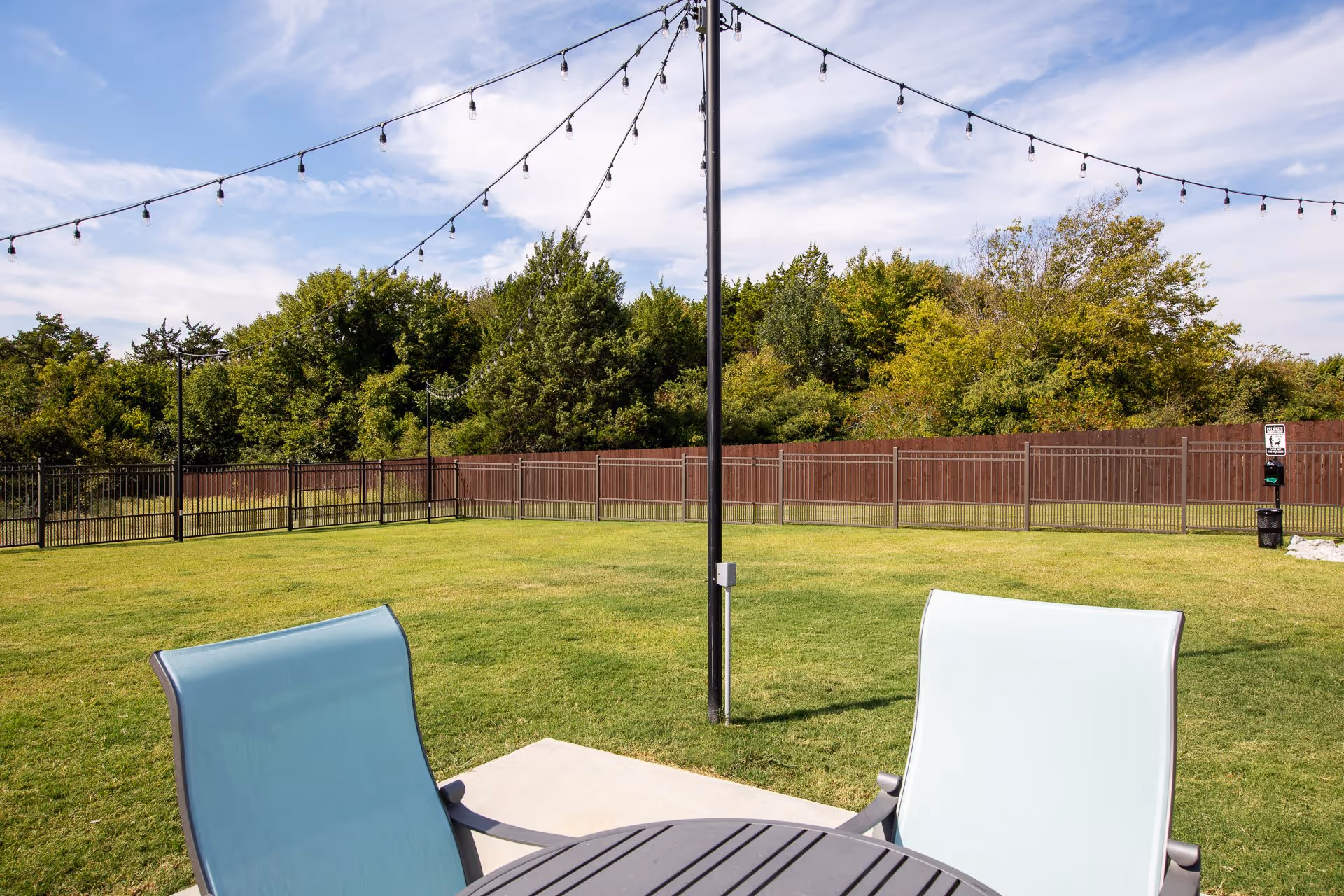 Outdoor seating area with two light blue chairs and a round table on a concrete patio, overlooking a large grassy fenced yard with string lights hanging overhead and trees in the background under a partly cloudy sky.