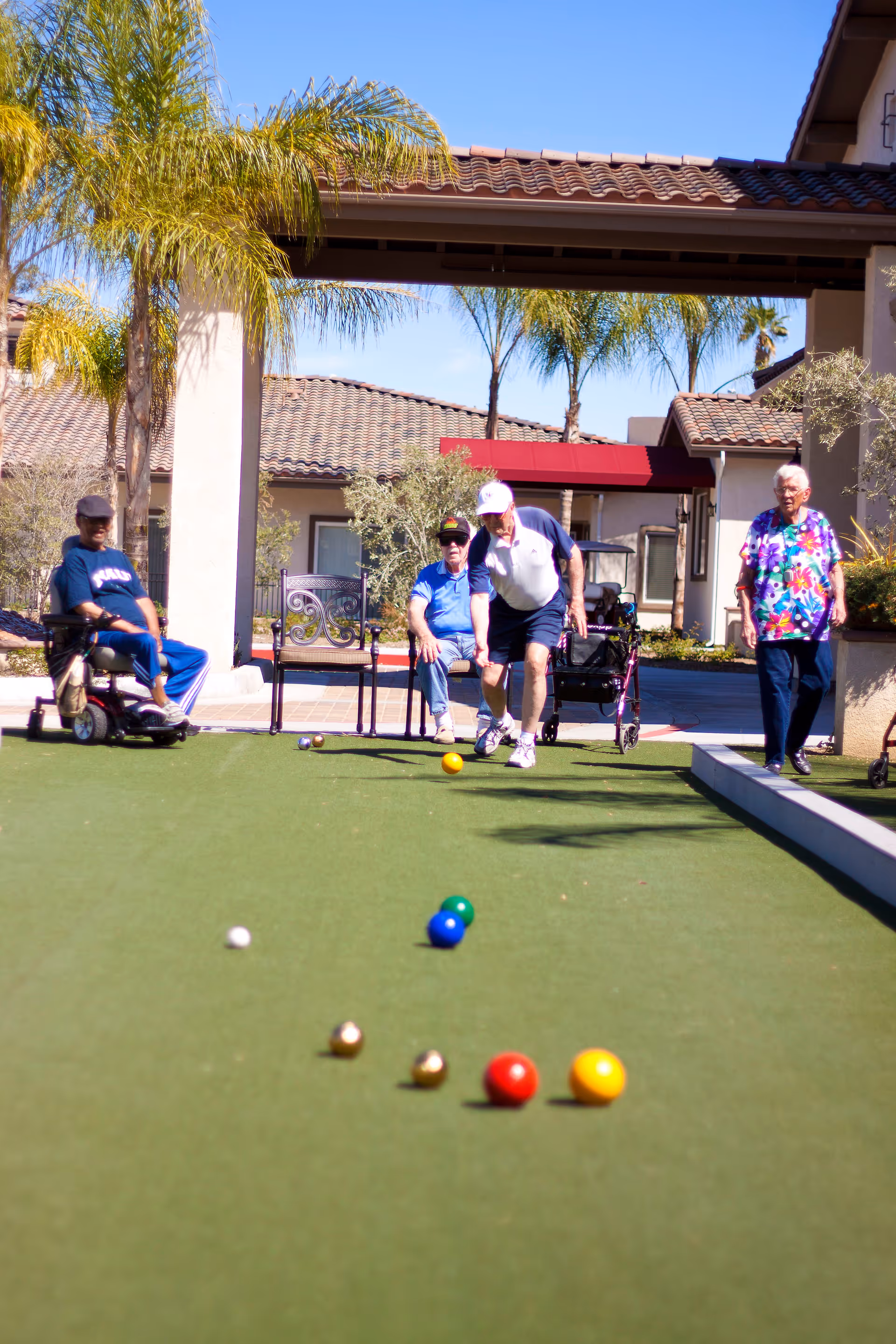 Four elderly people enjoying a game of bocce ball on a green court outdoors at a senior living facility with palm trees and tiled roof buildings in the background.