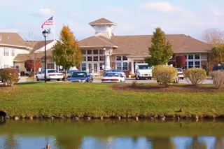 Exterior view of a senior living facility building named The Addison of Park Crescent with a parking lot in front, several cars parked, trees, and a pond reflecting the building and sky.
