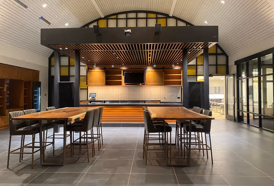 A modern dining area with two wooden tables and eight cushioned chairs arranged around them. The room features a high, curved ceiling with recessed lighting and a large counter area with wooden cabinets and a mounted TV screen in the background. There are large windows and glass doors on the right side, and additional cabinetry on the left.
