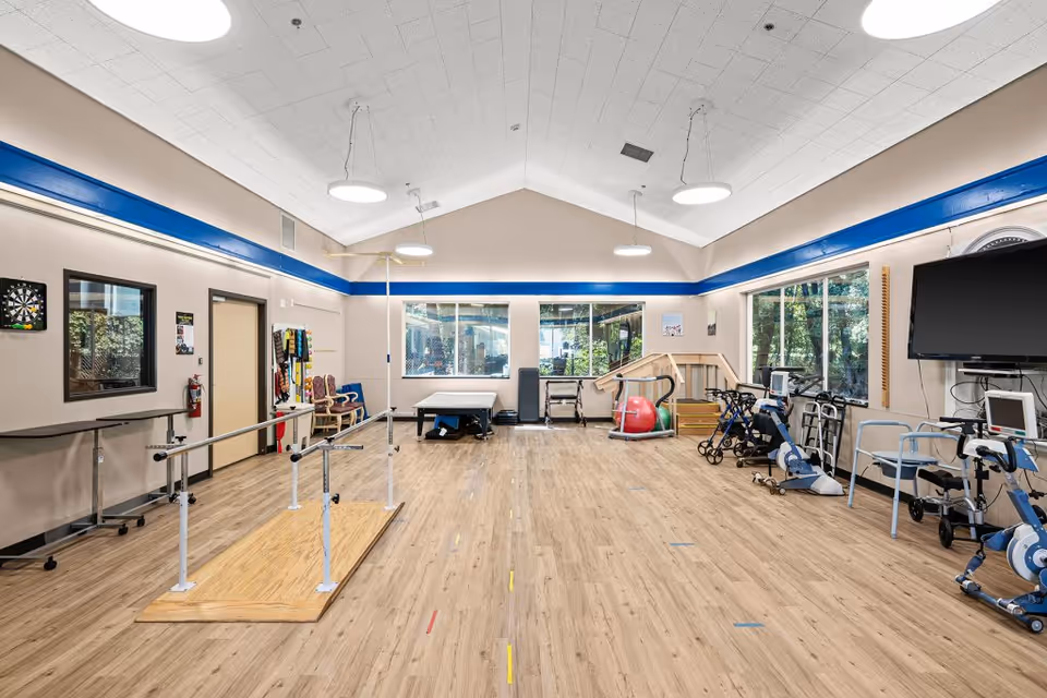 A spacious rehabilitation or physical therapy room with wooden flooring, parallel bars for walking practice, exercise equipment including stationary bikes, walkers, therapy balls, and a large TV mounted on the wall. The room has large windows letting in natural light and a high ceiling with circular hanging lights.