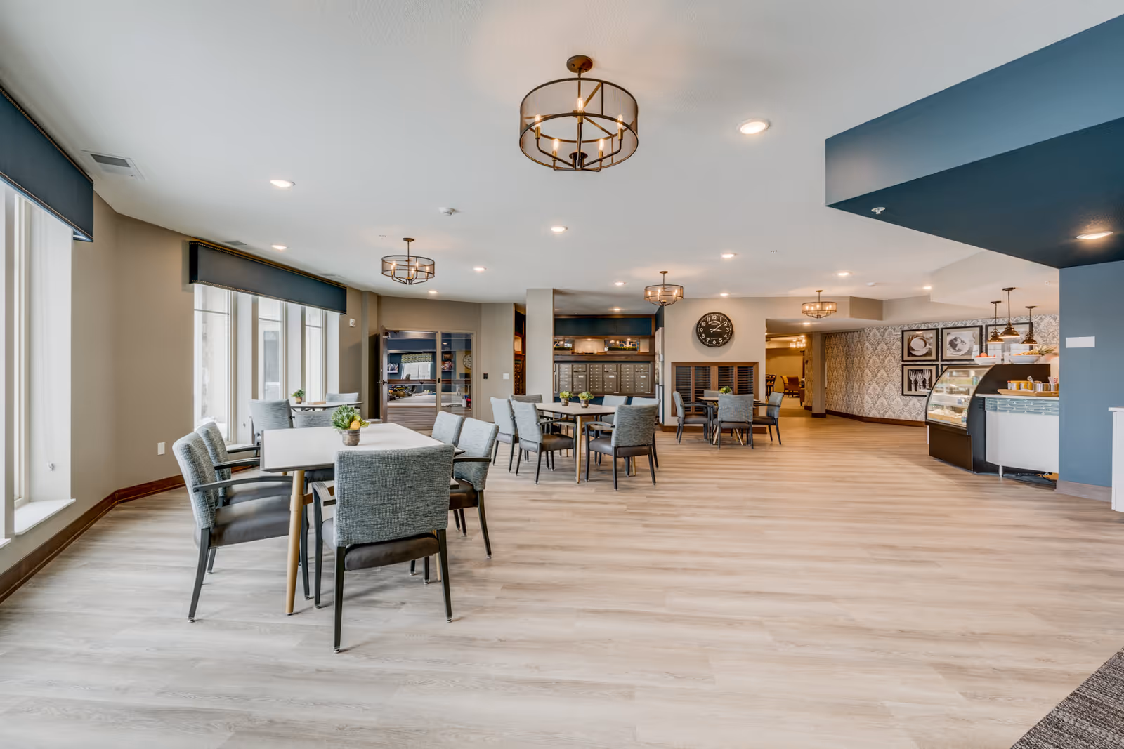 A spacious and well-lit dining area in a senior living facility with multiple tables and chairs arranged neatly. The room features large windows with dark blinds, modern light fixtures on the ceiling, and light wood flooring. In the background, there is a small café or food service counter with display cases and pendant lights, along with decorative wall art and a clock.