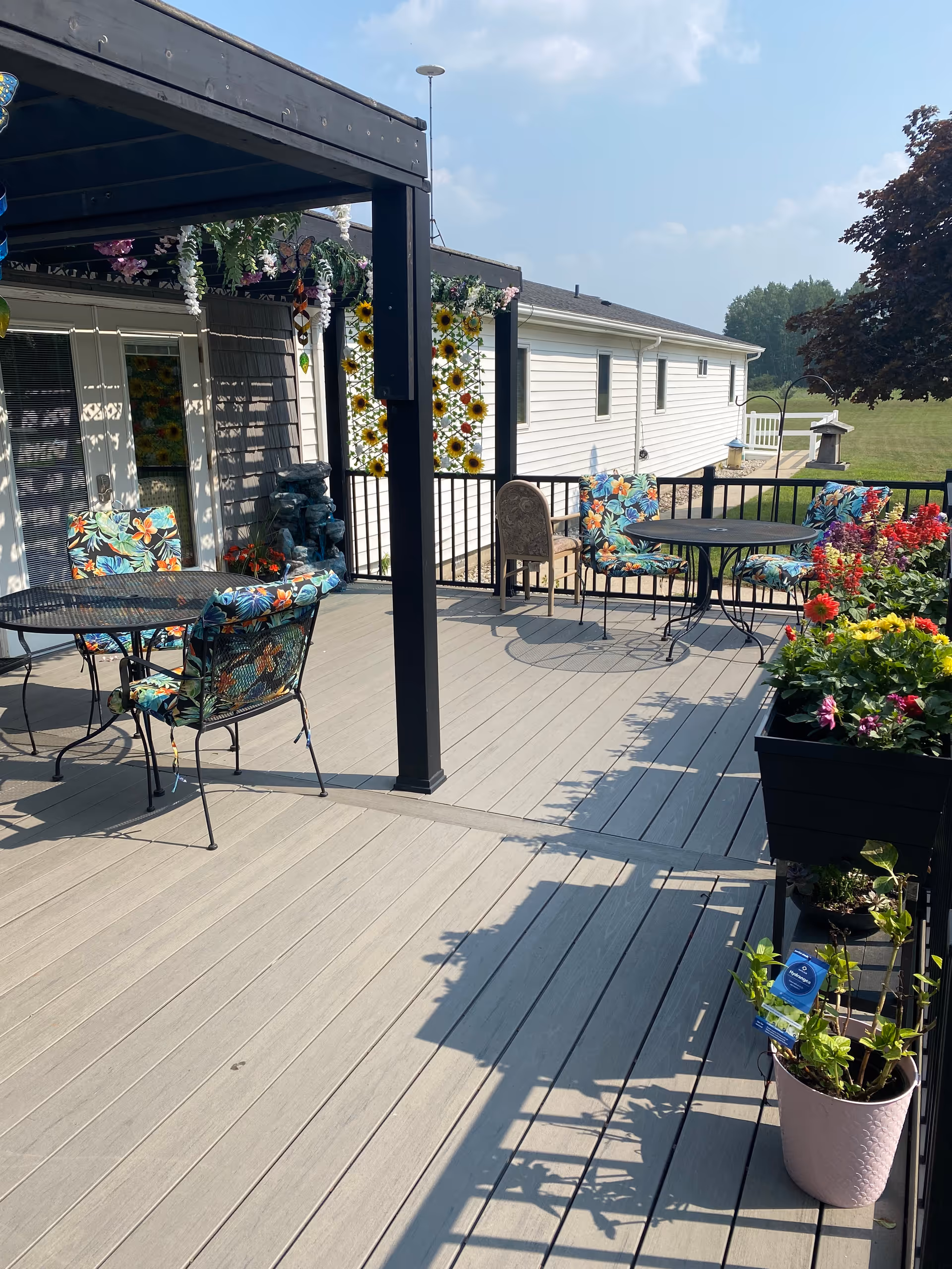 Outdoor patio area with a wooden deck featuring two round metal tables and several chairs with colorful floral cushions. There is a black pergola structure with hanging plants and decorations. Flower boxes with vibrant flowers and potted plants line the railing. A white building and green lawn are visible in the background under a partly cloudy sky.
