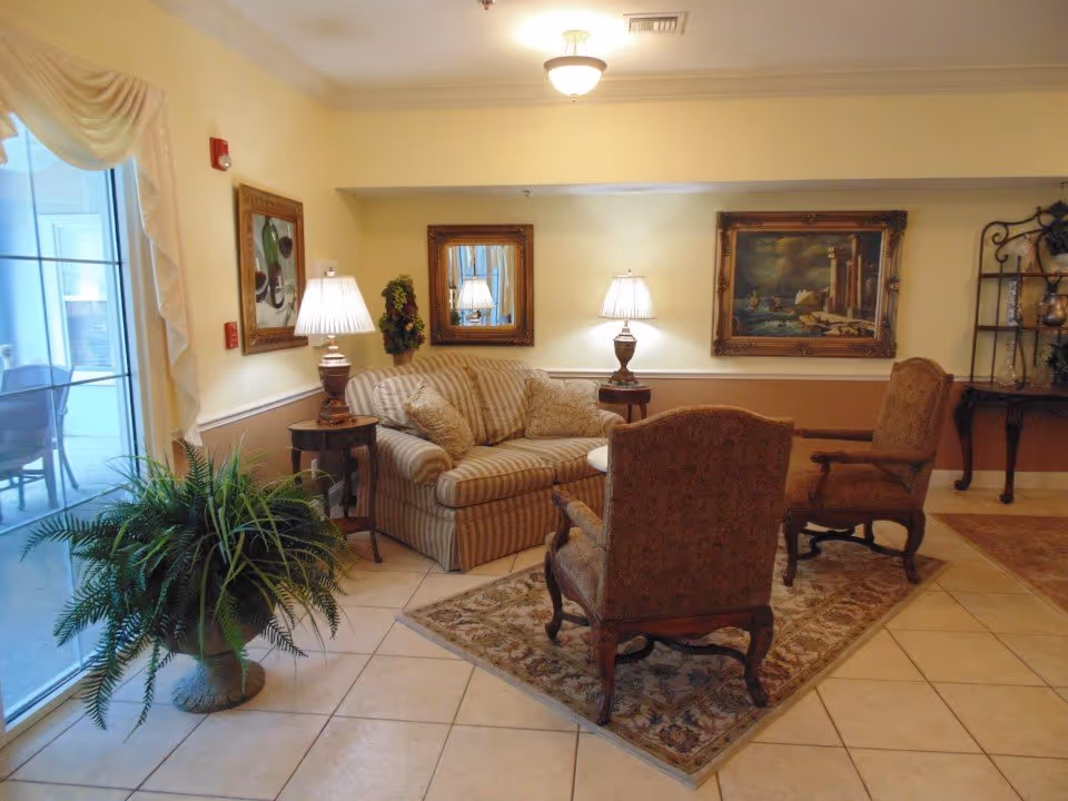 A cozy sitting area with a striped sofa, two armchairs, lamps, framed artwork, and a large potted plant in a tiled common room.