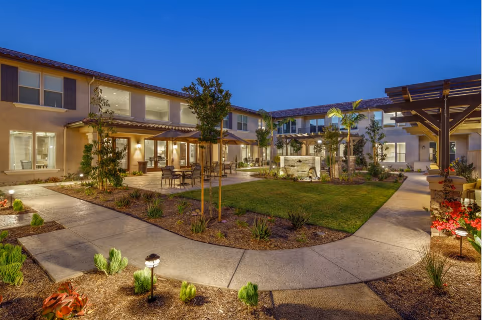 Evening view of an outdoor courtyard at Oakmont of Riverpark featuring a well-lit pathway, green lawn, small trees, patio tables with umbrellas, and a water fountain surrounded by a two-story building.