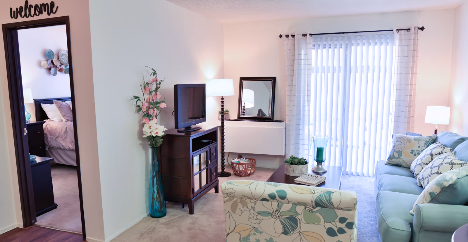 Bright living room with a sofa, patterned armchair, TV on a cabinet, coffee table, and sliding glass doors, with a bedroom visible through an open doorway.