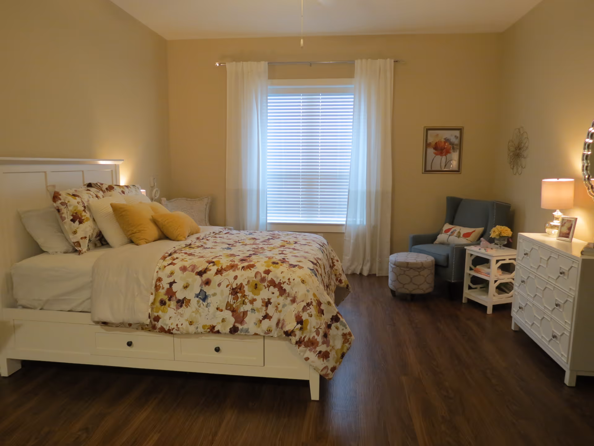 A cozy bedroom with a white bed frame and floral bedding, several pillows including yellow ones, a window with white curtains and blinds, a blue armchair with a decorative pillow, a small round ottoman, a white side table with flowers and books, a white dresser with a lamp and framed photo, and wall decorations including a flower picture and a metal wall art piece.