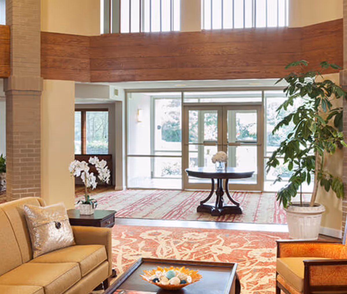 Sunny lobby with upholstered seating, a coffee table, potted plants, and glass entrance doors.