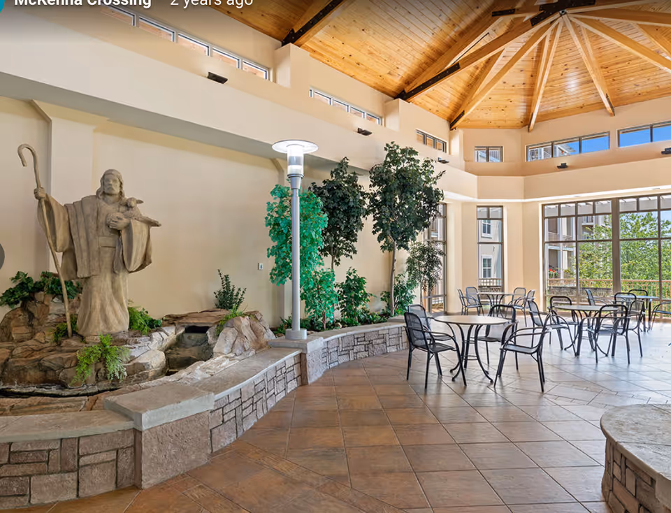 Spacious indoor common area with a high wooden ceiling and large windows letting in natural light. The room features several round tables with black metal chairs arranged for seating. On the left side, there is a stone statue of a shepherd holding a staff and a lamb, surrounded by greenery and a small water feature. The floor is tiled with a warm-toned pattern.