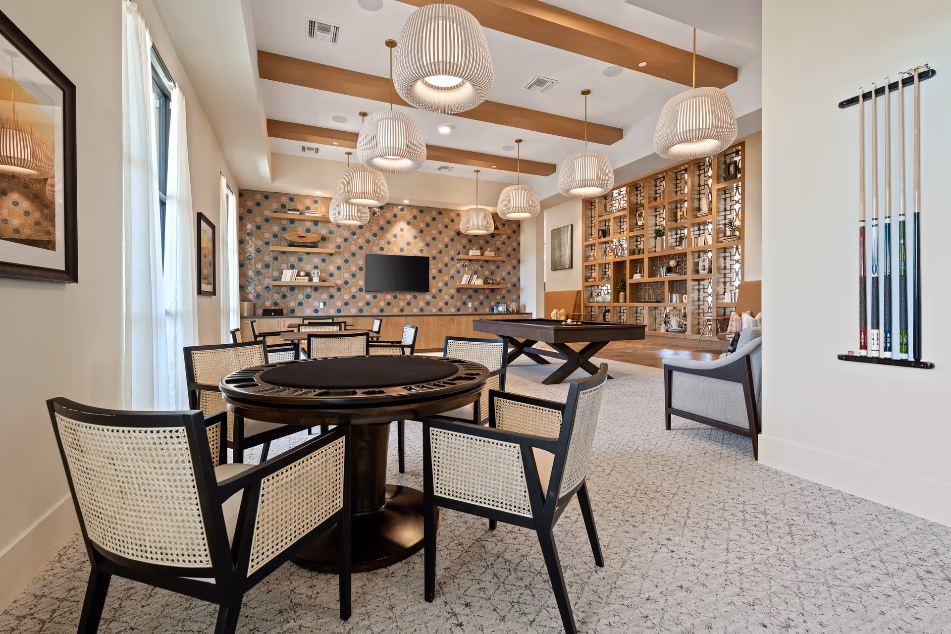 A spacious and well-lit game room featuring a round card table with six chairs, a pool table, and a wall-mounted TV. The room has modern pendant lights hanging from a ceiling with wooden beams, a patterned accent wall with shelves holding decorative items, and a large wooden shelving unit with various decor pieces. Pool cues are mounted on the wall to the right.