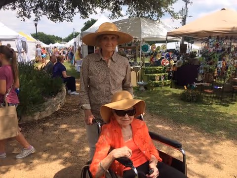 An elderly woman wearing a large sunhat and sunglasses sits in a wheelchair, while an elderly man wearing a straw hat stands behind her pushing the wheelchair. They are outdoors at a market or fair with tents and people in the background.