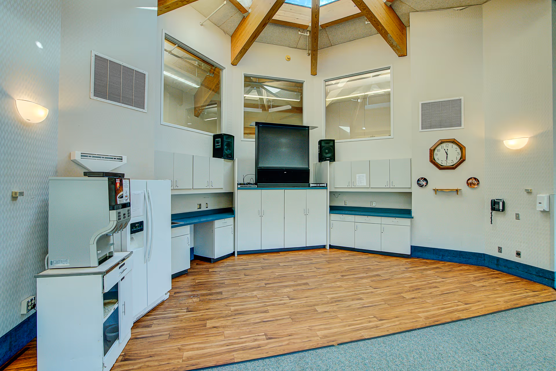 A spacious room with wooden flooring and white walls featuring built-in cabinets and countertops along two walls. There is a large TV mounted in the center with two speakers on either side. A white refrigerator and a coffee machine are positioned on the left side. The ceiling has wooden beams and skylights, and there is a wall clock and two decorative plates on the right wall.