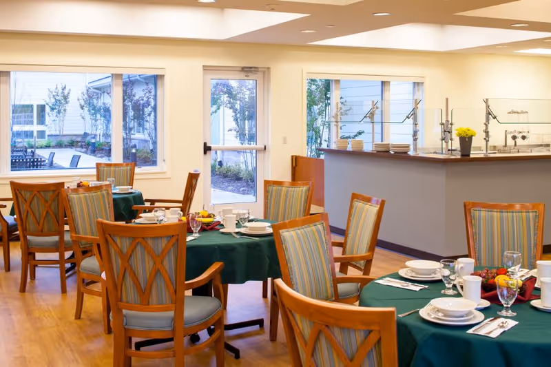 A bright dining room with several round tables covered with green tablecloths, set with white plates, cups, glasses, and silverware. Wooden chairs with striped cushions surround the tables. Large windows and a glass door provide a view of an outdoor garden area. A serving counter with plates and a small yellow flower pot is visible in the background.