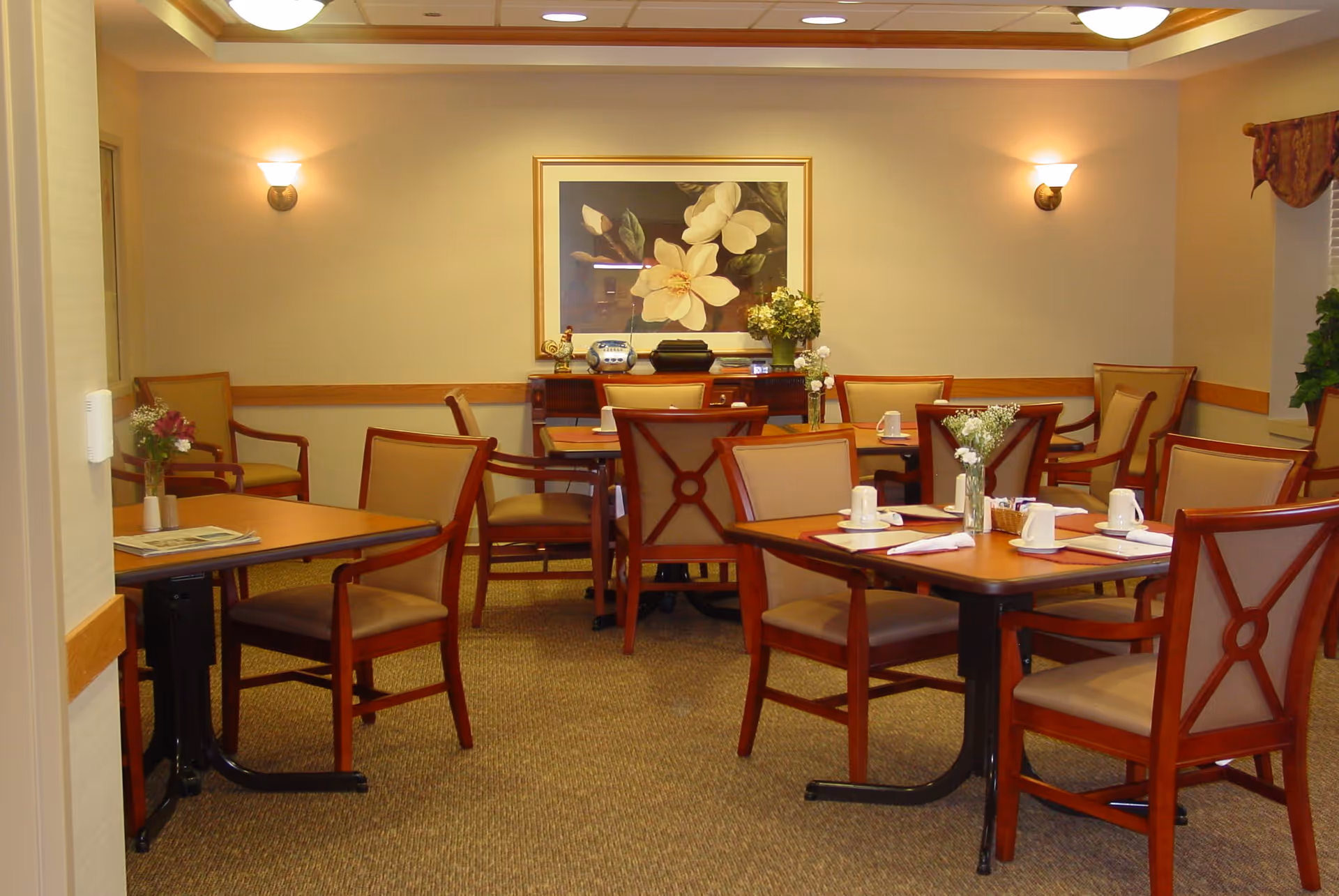A dining room with several wooden tables and chairs arranged neatly. Each table has placemats, napkins, cups, and small flower vases. The room has beige walls with two wall sconces providing warm lighting. A large framed floral painting hangs on the far wall above a sideboard with decorative items and flowers.