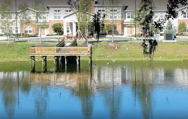 A wooden viewing pier extends over a pond with reflections and a multi-story residential building behind it.