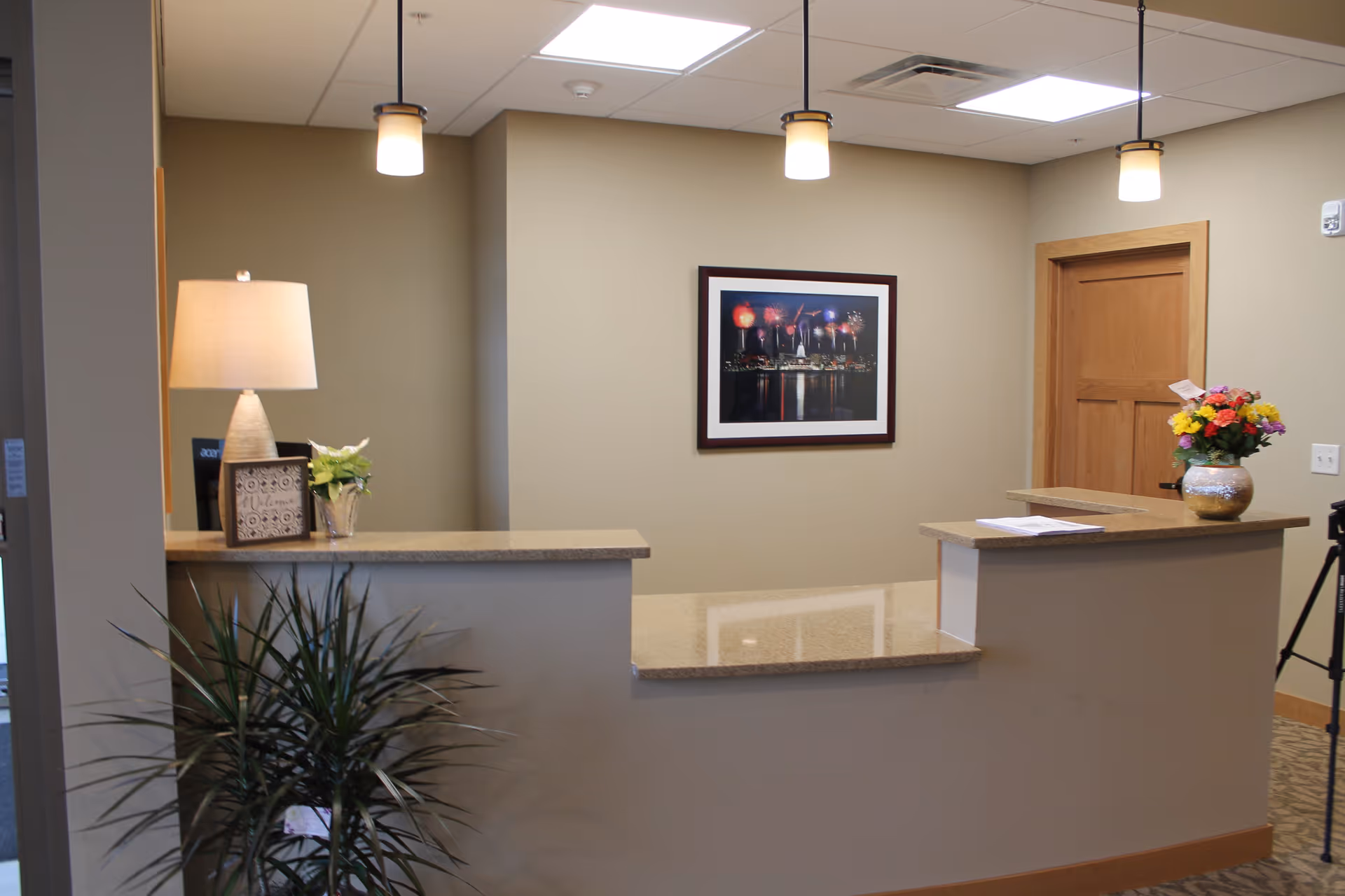Reception desk area in Oak Park Place Nakoma with a lamp, potted plants, a framed picture of fireworks on the wall, and a vase of colorful flowers on the counter. The space is well-lit with ceiling lights and has a wooden door in the background.
