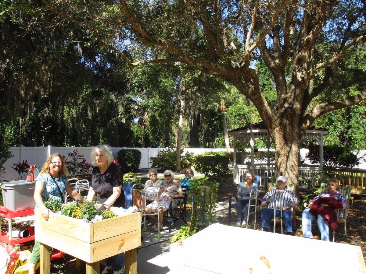 A group of elderly people sitting outdoors in a garden area under large trees, with two women standing near a wooden planter box filled with plants. The setting includes a white fence, greenery, and a gazebo in the background.