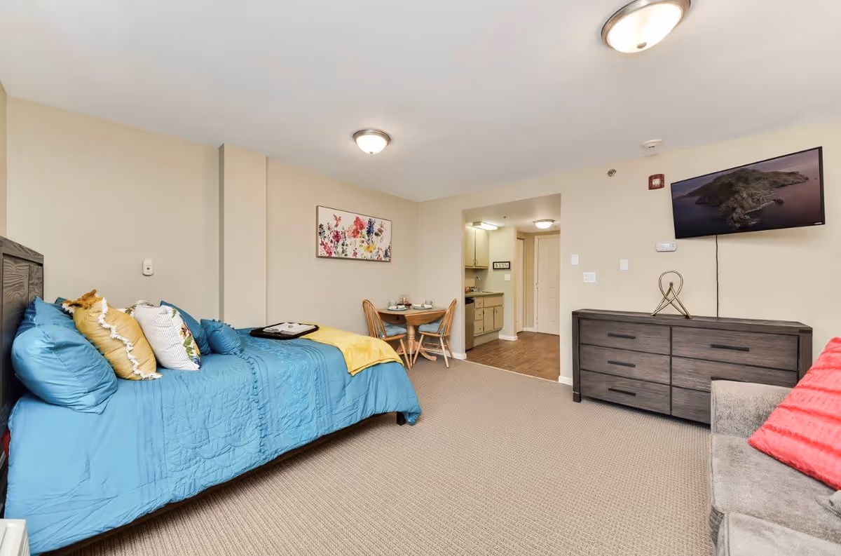 A cozy living space in an assisted living facility featuring a bed with blue bedding and multiple pillows, a small round dining table with two chairs, a dark wooden dresser with a decorative item on top, a wall-mounted flat-screen TV, and a gray couch with a red pillow. The room has beige walls, carpeted flooring, and ceiling lights. An open doorway leads to a small kitchen area.