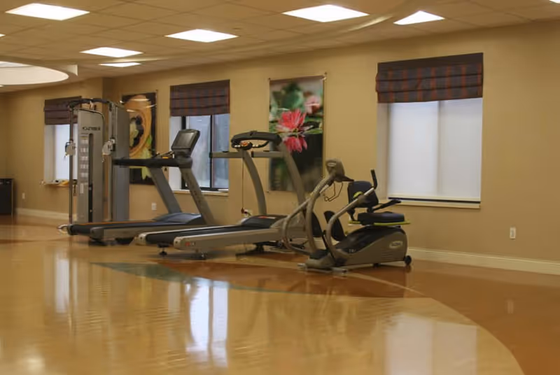 A fitness room with exercise equipment including two treadmills, a recumbent bike, and a multi-function weight machine. The room has a polished wooden floor, beige walls, three windows with brown plaid valances, and a floral wall decoration.