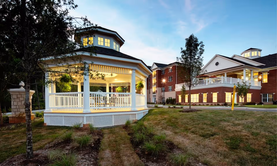 A lit white gazebo on a lawn in front of a multi-story brick senior living building at dusk.