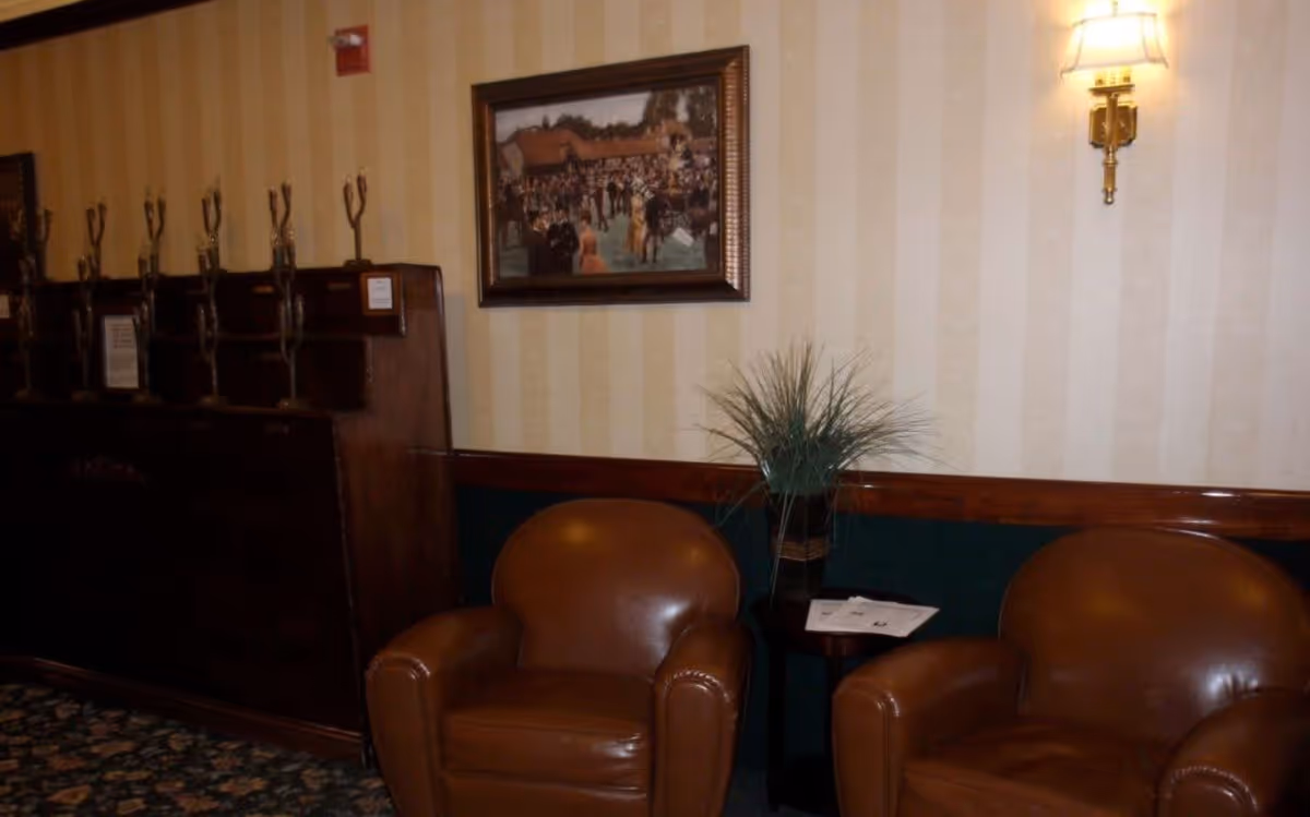 A cozy seating area with two brown leather armchairs separated by a small wooden table holding a plant and some papers. Behind the chairs is a wall with striped wallpaper, a framed painting, and a wall-mounted light fixture. To the left, there is a wooden coat rack with multiple hooks.