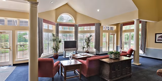 A spacious living room area with large windows allowing natural light to fill the space. The room features red upholstered chairs and sofas arranged around a central coffee table with decorative items. There are two tall potted plants near the windows, a television on a stand, and a wooden side table with a plant on it. The walls are painted in warm tones with white trim, and the ceiling is vaulted with recessed lighting.