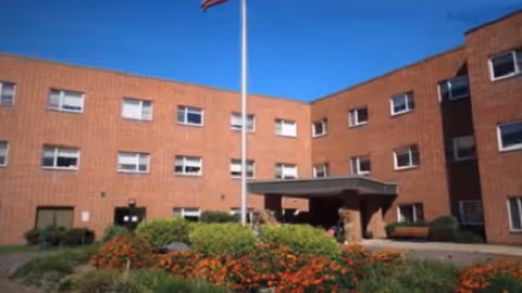 Exterior view of a three-story brick nursing and rehabilitation center building with multiple windows, a covered entrance, a flagpole with an American flag, and landscaped flower beds in front.