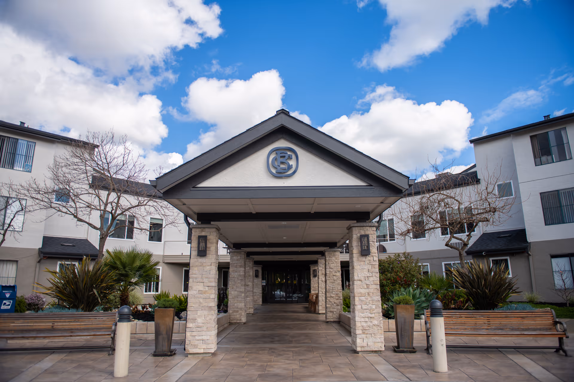 Entrance of a senior living facility with a covered walkway supported by stone pillars, benches on either side, and landscaped plants. The building is white with multiple windows and a blue sky with clouds in the background.