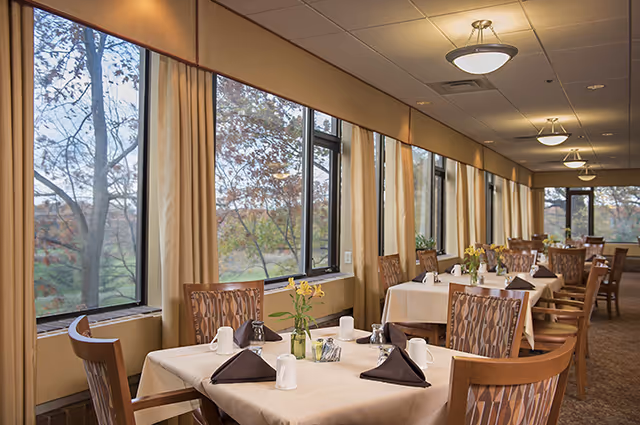 Dining area with several tables covered with beige tablecloths, each set with white mugs, silverware, salt and pepper shakers, and small vases with yellow flowers. Large windows with beige curtains line one side, allowing natural light and a view of trees outside.