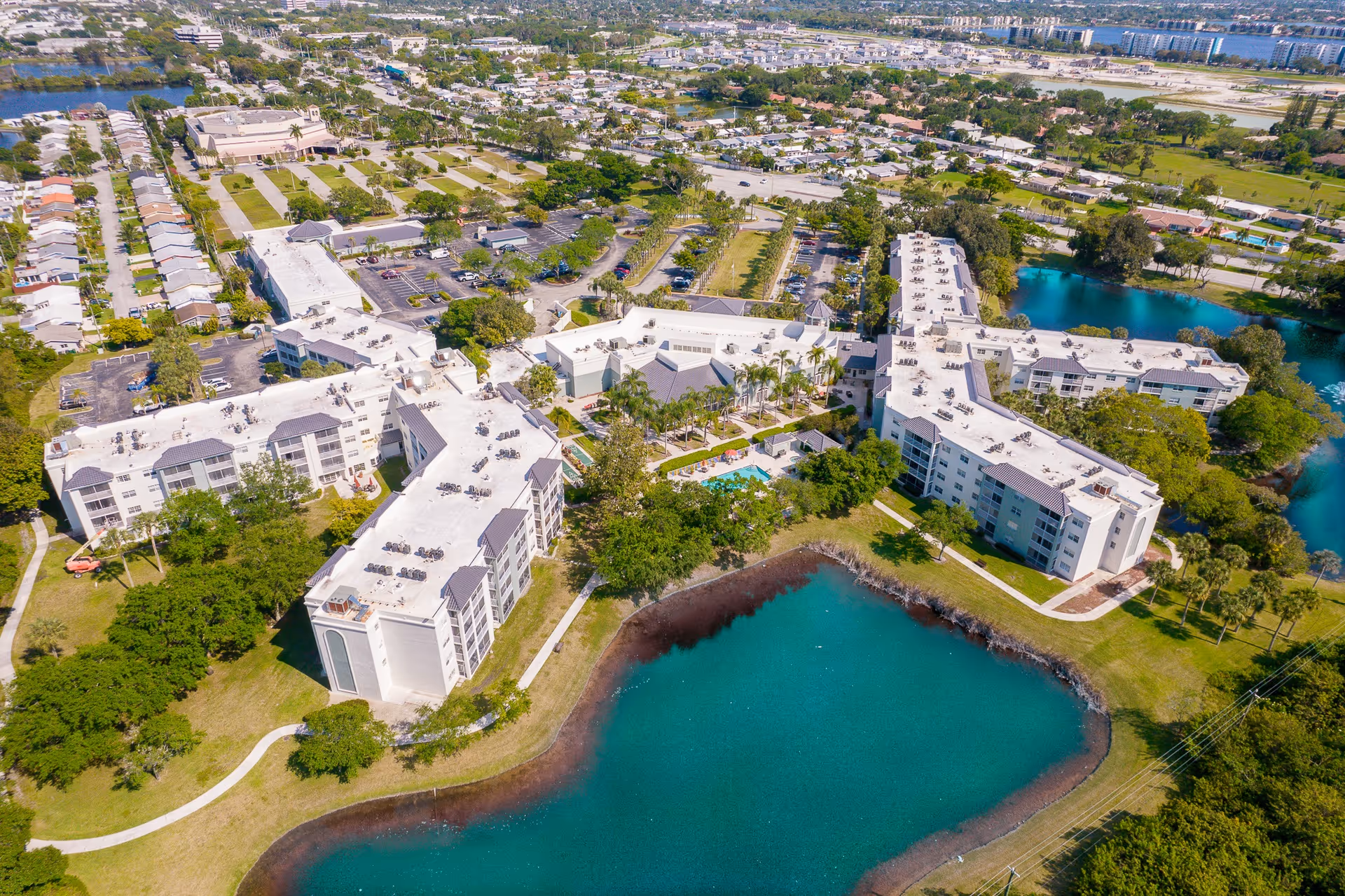 Aerial view of a large senior living facility named Oasis Living Quarters, featuring multiple connected white buildings with flat roofs surrounding a central courtyard with a swimming pool. The facility is bordered by a pond and landscaped green areas, with parking lots and residential neighborhoods visible in the background.