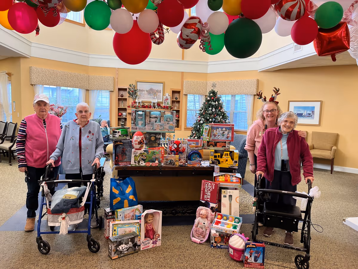 Four elderly women standing indoors in a decorated room with Christmas balloons hanging from the ceiling. Two women are using walkers. In front of them is a table filled with various toys and gifts, with more toys placed on the floor. A decorated Christmas tree and festive decorations are visible in the background.