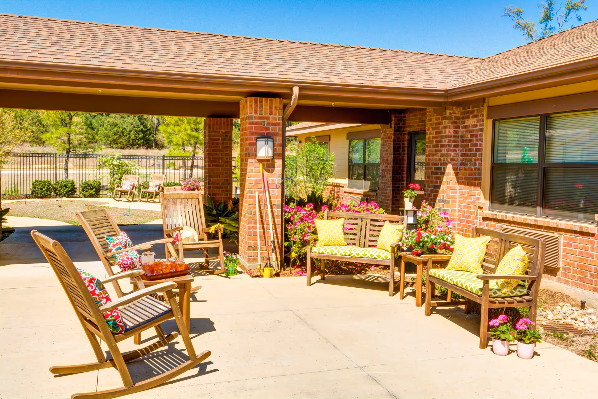 Sunlit outdoor patio at a brick memory care facility with wooden rocking chairs, benches with colorful pillows, and potted flowers.