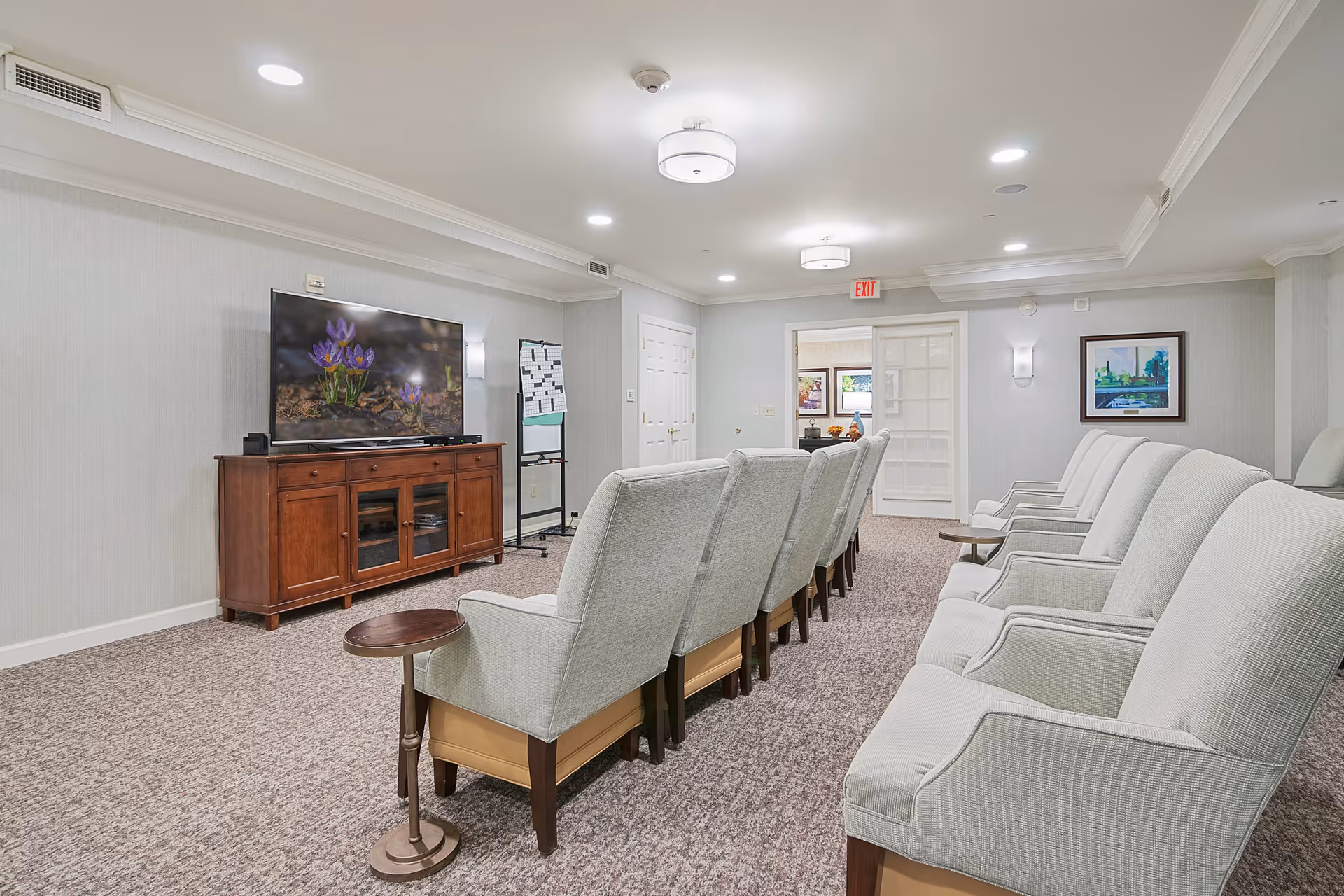 A cozy common room with two rows of light gray upholstered armchairs facing a large flat-screen TV mounted on a wooden cabinet. The room has beige carpet, light gray walls, recessed ceiling lights, and framed artwork on the walls. A whiteboard on a stand is visible near the TV, and a door with glass panels leads to another room.
