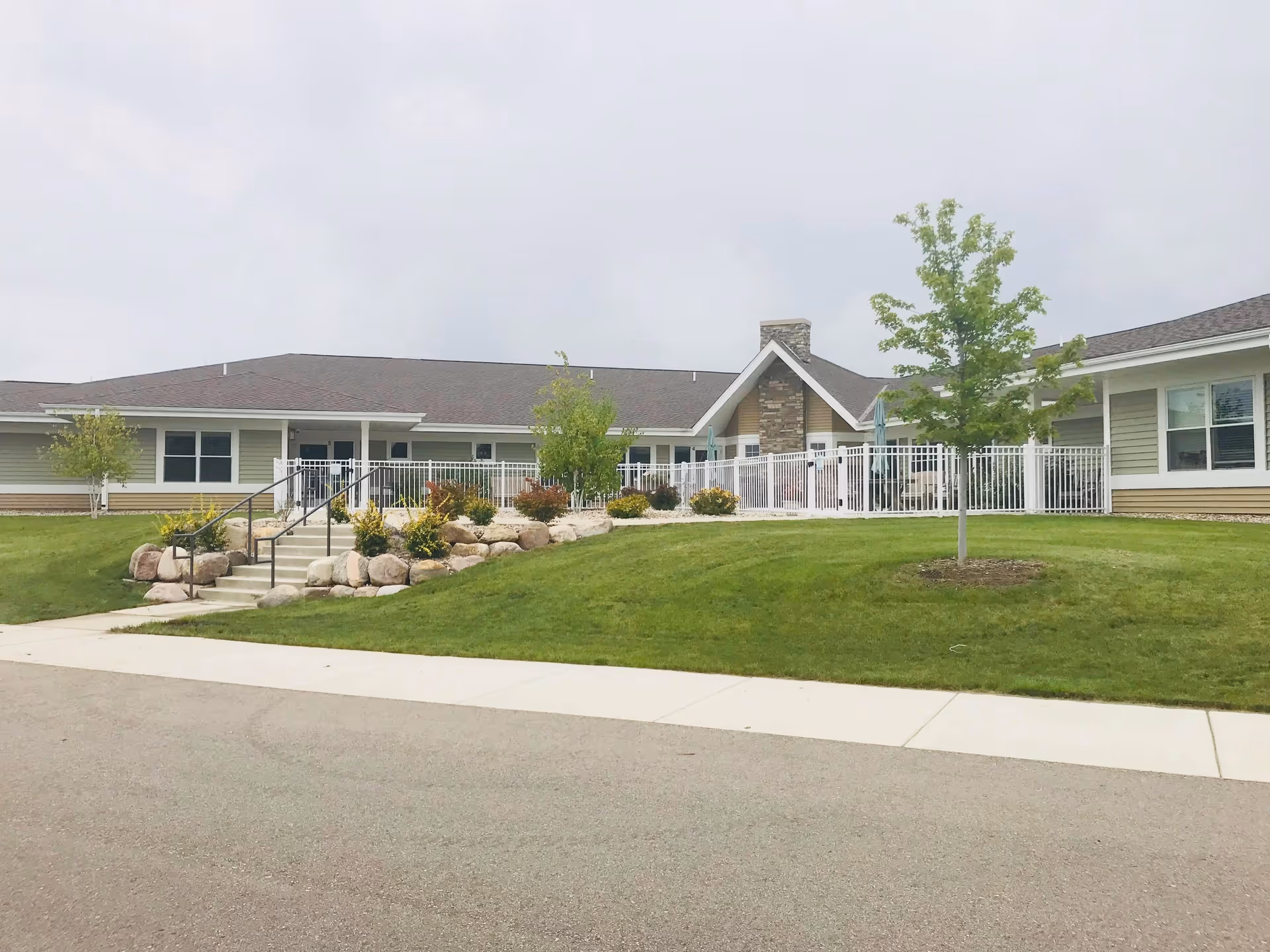 Exterior view of Granite Falls Health Care Center showing a single-story building with a sloped roof, stone chimney, white railing, and a landscaped lawn with trees and shrubs.