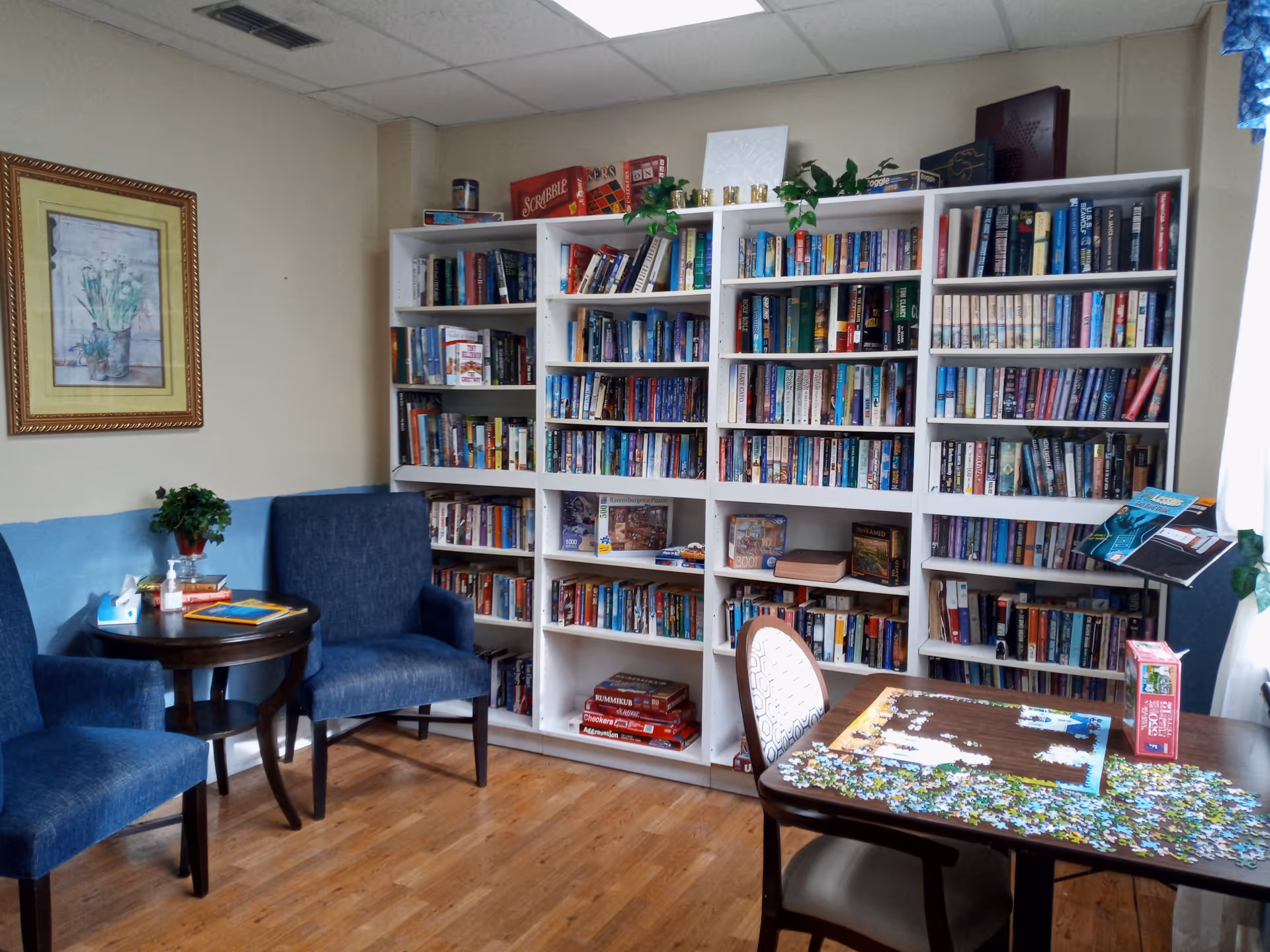 A cozy reading and activity room with two blue upholstered chairs around a small round wooden table holding books, tissues, and hand sanitizer. A large white bookshelf filled with books and board games stands against the wall. A wooden table with a partially completed jigsaw puzzle and a chair is in the foreground. The room has light-colored walls, a framed floral painting, and a window with blue curtains letting in natural light.