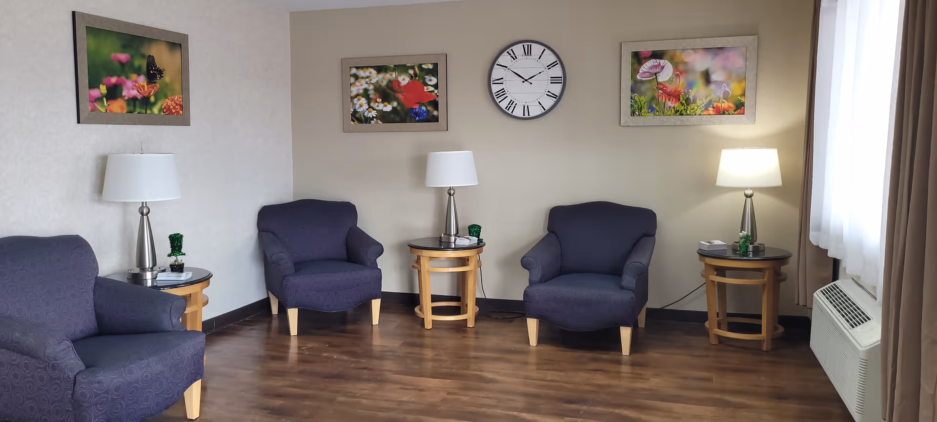A bright common sitting room with four upholstered armchairs arranged around side tables, table lamps, a wall clock and floral artwork.