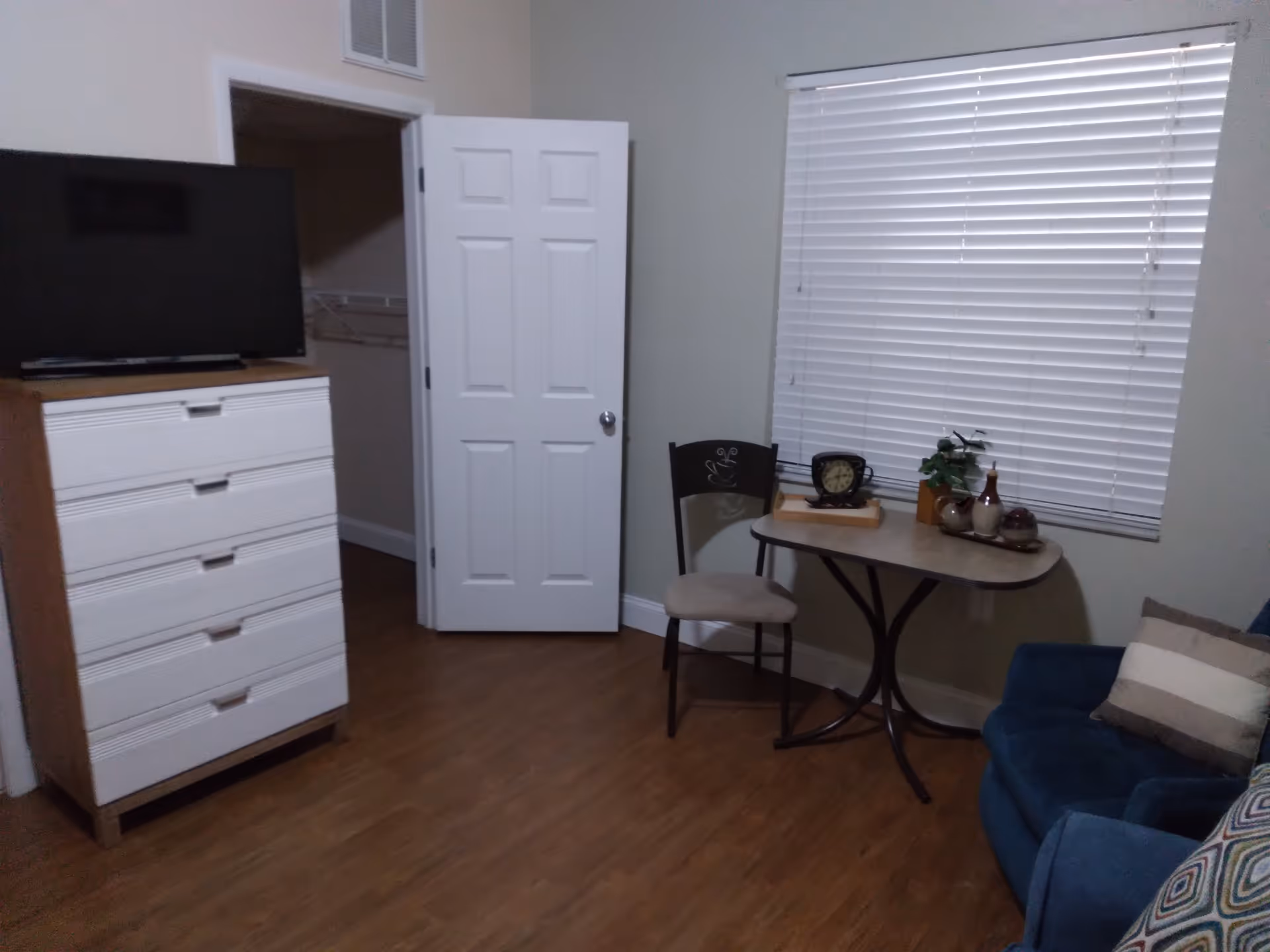 Interior of a senior living room with a wooden floor, a white chest of drawers with a TV on top, an open door revealing a closet, a small table with a chair, a clock, decorative items, and a blue armchair with a striped pillow near a window with closed blinds.
