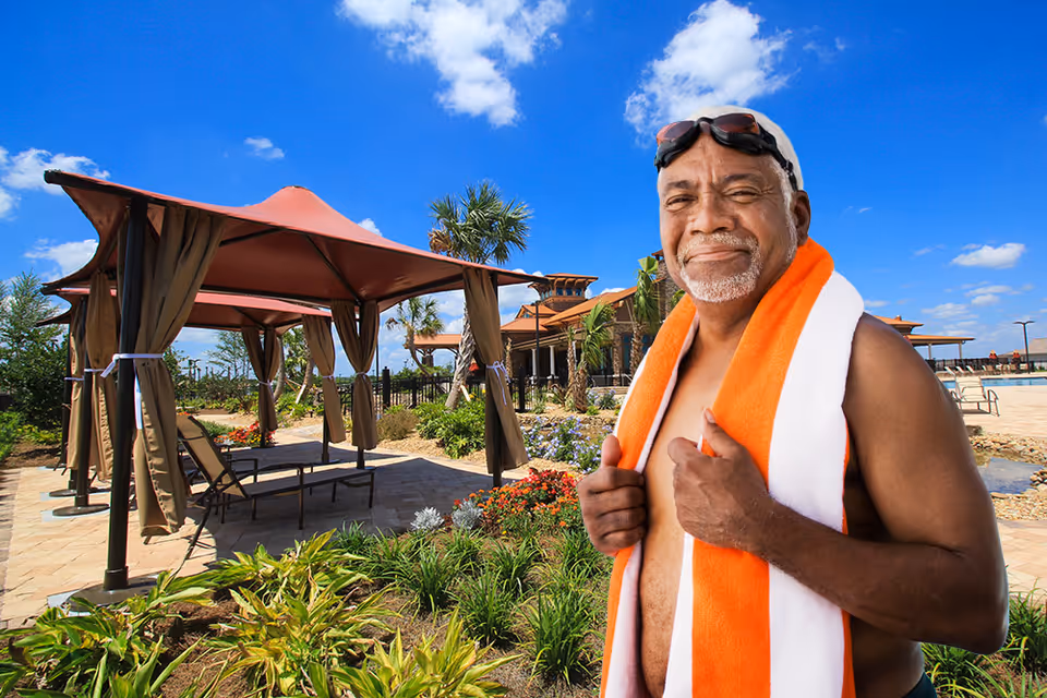 A smiling man with swim goggles and an orange-and-white towel stands by poolside cabanas and landscaped gardens under a bright blue sky.