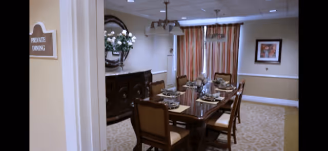 A private dining room with a rectangular wooden table set for six people. The table has place settings with plates, glasses, and napkins. There are six cushioned chairs around the table. The room features a sideboard with a decorative mirror above it and a vase of white flowers. A window with striped curtains and a framed picture on the wall are visible. A sign on the wall near the entrance reads 'PRIVATE DINING'.