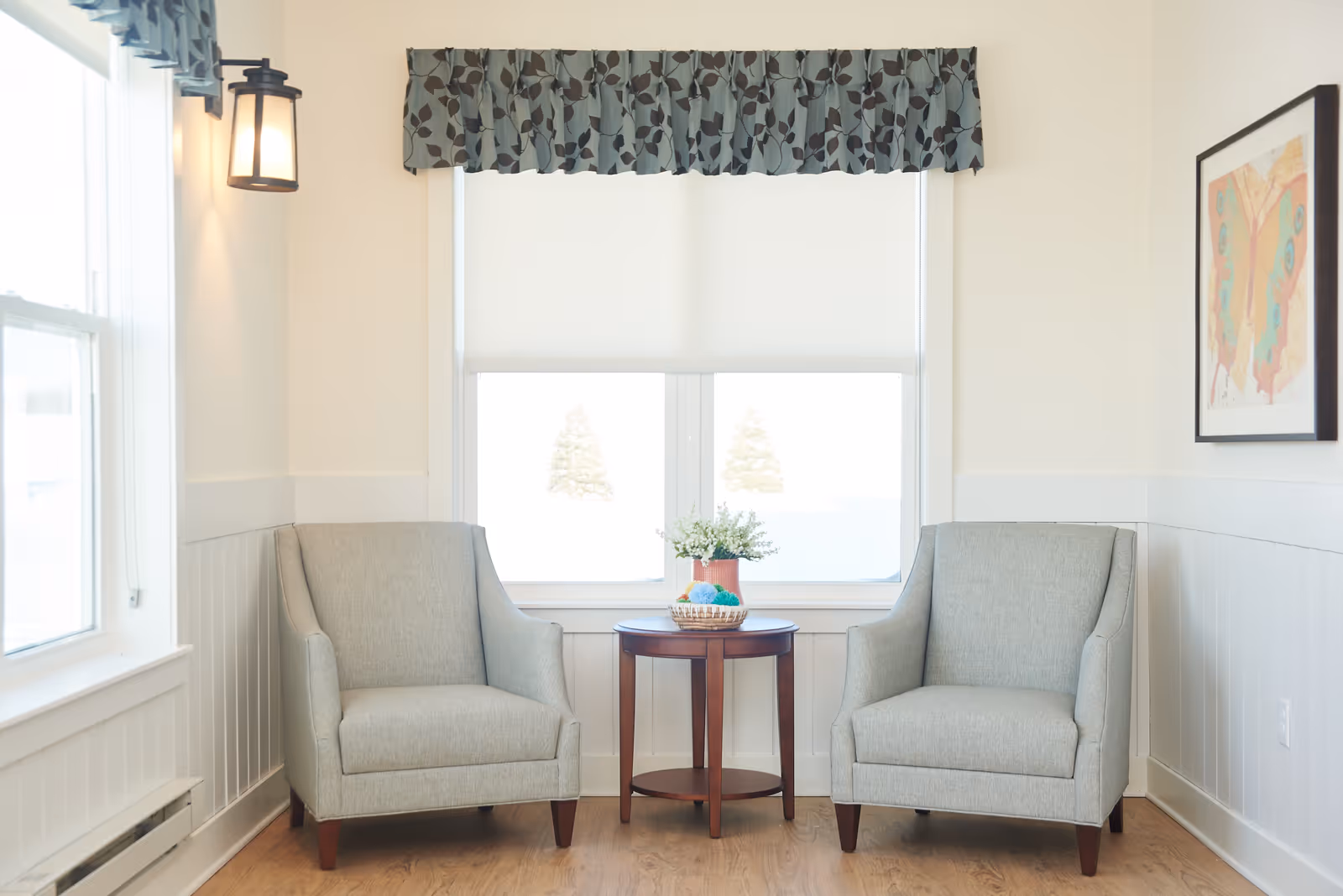 A cozy sitting area with two light gray armchairs positioned on either side of a small round wooden table. The table holds a small plant and a basket with decorative balls. Behind the chairs is a window with a white roller shade and a patterned valance. The walls are light-colored with white wainscoting, and there is a framed butterfly artwork on the right wall. A lantern-style wall light is mounted on the left wall.