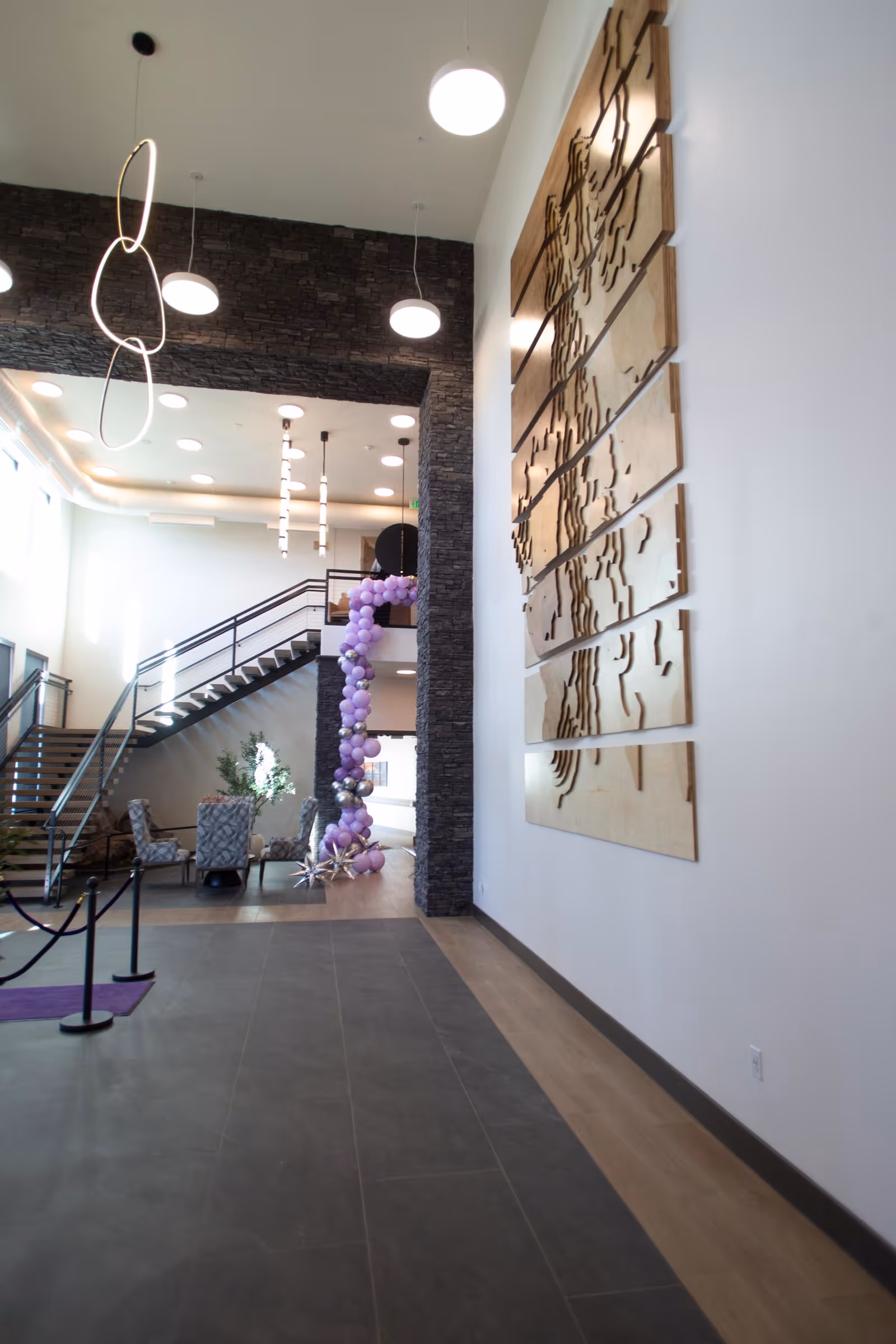 Spacious modern lobby with high ceilings, a staircase, seating area, large wooden wall art, and a column of purple balloons.