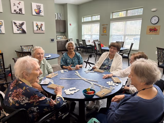 A group of six elderly women sitting around a round table playing dominoes in a bright room with large windows and wall decorations including pictures of dogs and a clock.