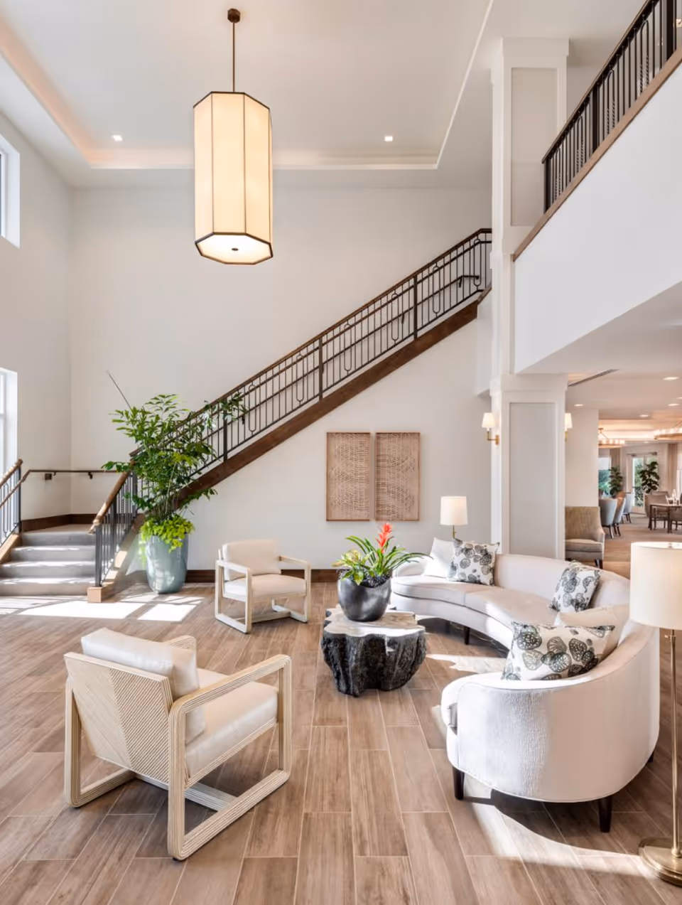 A bright and spacious living area with a curved white sofa adorned with patterned pillows, two modern armchairs, a unique tree stump coffee table with a potted plant, and a large hanging light fixture. A staircase with a dark wooden handrail and metal balusters leads to an upper floor. The room features light wood flooring, white walls, and large windows allowing natural light to fill the space.