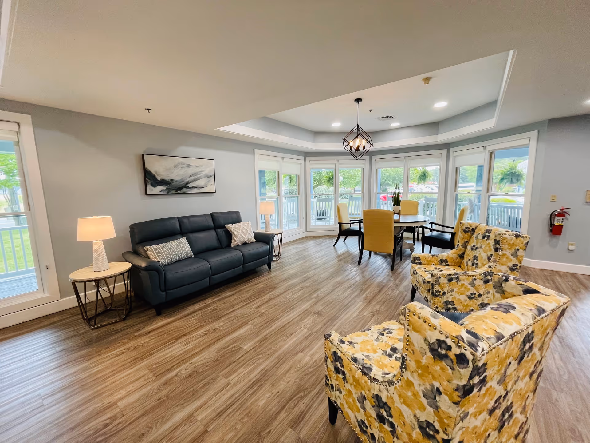A bright and spacious common area in an assisted living facility featuring a black leather sofa with two patterned pillows, two yellow floral armchairs, a round dining table with six yellow chairs, large windows with white blinds letting in natural light, a modern geometric pendant light, and wood-look flooring.
