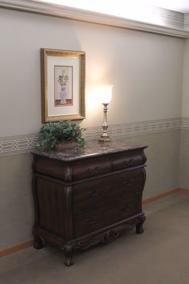 Ornate wooden dresser with a marble top topped by a lamp and potted plant beneath a framed floral picture on a beige hallway wall.