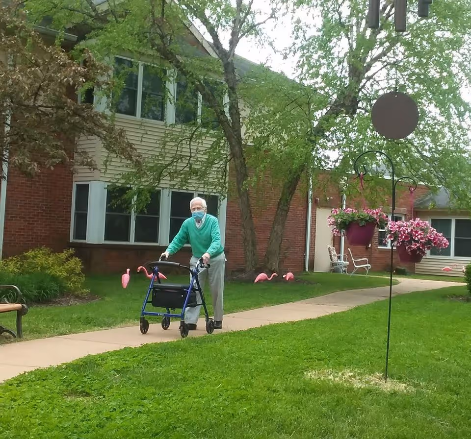 An elderly person wearing a green sweater and a face mask is walking with a blue walker on a paved path outside a brick building. The area is grassy with trees, pink flamingo lawn ornaments, hanging flower baskets, and outdoor benches.