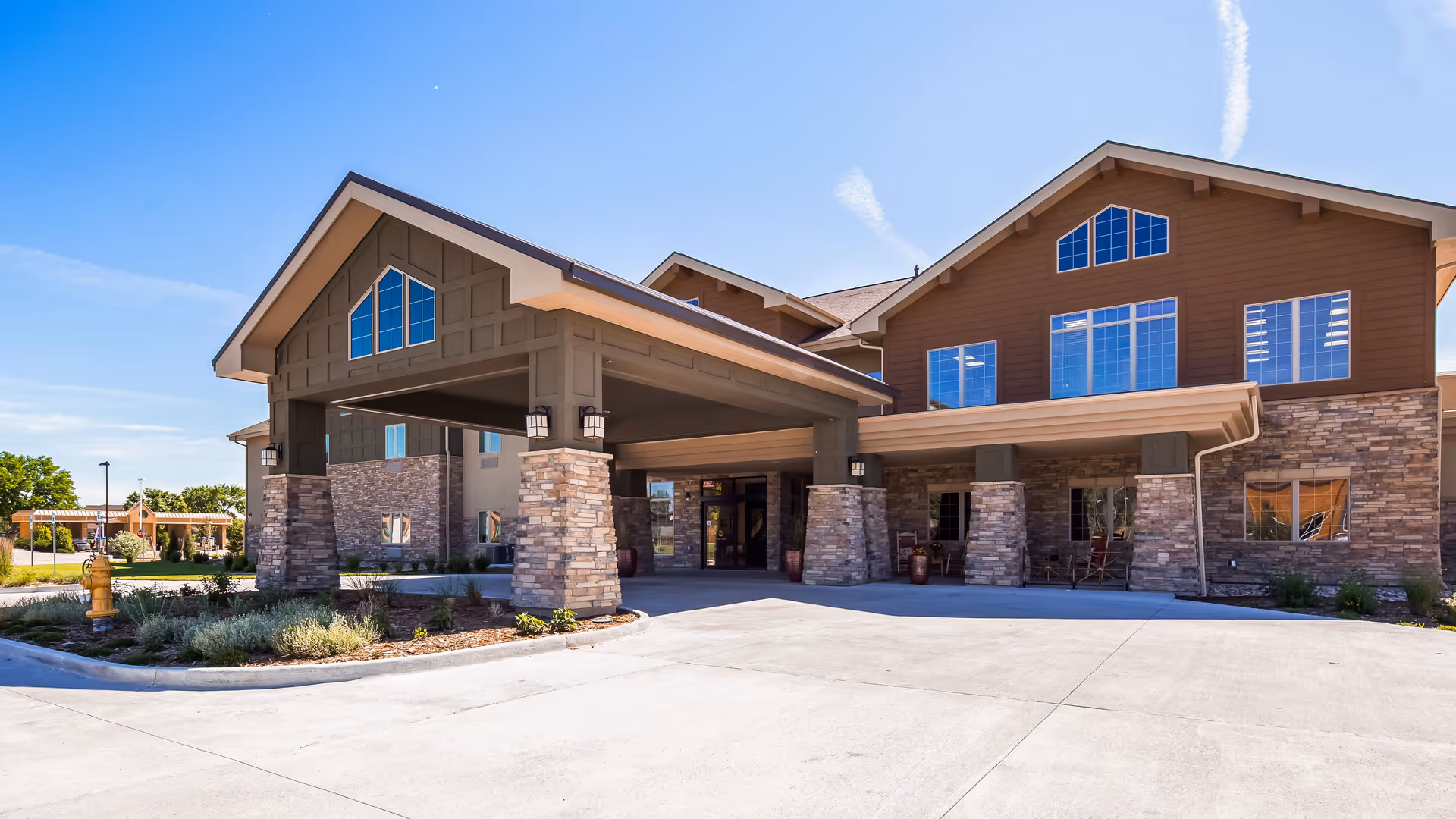 Exterior view of the Legend of Broomfield senior living facility showing a large covered entrance with stone pillars, brown siding, multiple windows, and a clear blue sky.